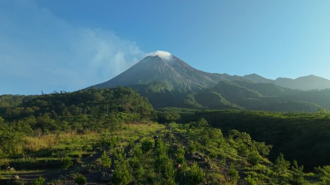 Aerial view of Mount Merapi with smoke rising from the crater above lush green hills and pine trees in Kecamatan Pakem, Daerah Istimewa Yogya, Indonesia.