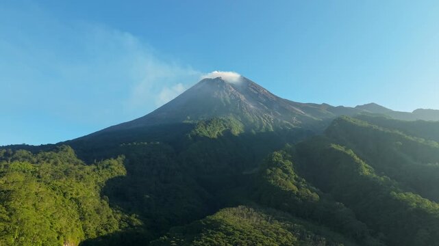 Aerial view of Mount Merapi volcano with smoke rising from the crater above lush green forest slopes in Kecamatan Pakem, Daerah Istimewa Yogya, Indonesia.
