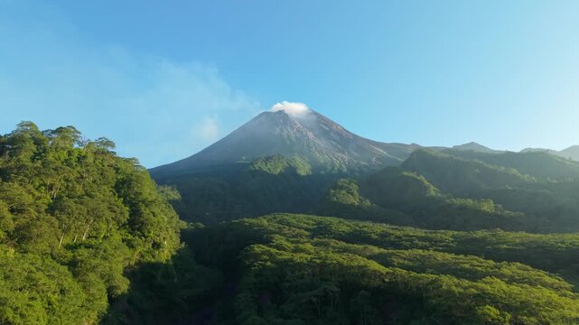 Aerial view of Kali Gendol River and Mount Merapi volcano with steam rising from the crater above dense tropical forest and rolling green hills in Kecamatan Pakem, Yogyakarta, Indonesia.