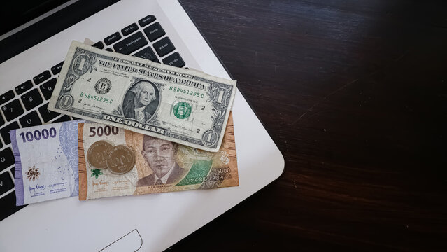 Comparison and exchange rate of US dollar and Indonesian rupiah banknotes, with a laptop in the background.