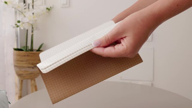 Hands holding open a small checkered notebook over a table, detailed view of paper texture and binding.