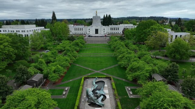 Oregon State Capitol building in Salem with iconic golden Pioneer statue atop Art Deco dome, surrounded by manicured green lawns and cherry blossoms. Drone backwards Wide aerial shot. Cloudy day in us