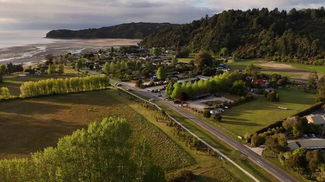 Marahau township and main street at sunset with lush forest mountain backdrop and view of tidal estuary in Abel Tasman National Park area, New Zealand. Aerial approaching flight.