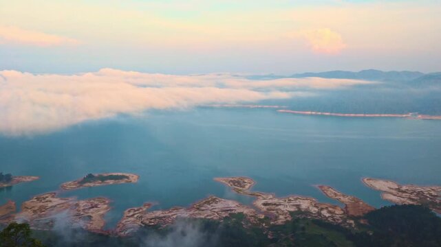 Cinematic morning pan over Tasik Pedu, Kedah. A thick bank of white mist rolls over turquoise lake waters and silhouetted tropical islands under a soft pastel dawn sky.