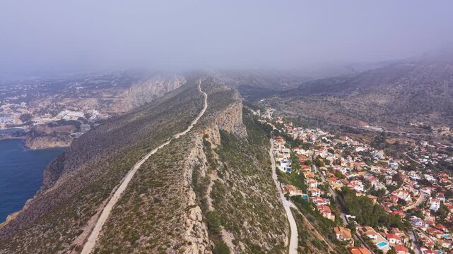Morro de Toix viewpoint overlooking Calpe, Spain, with rocky ridge, winding trail, coastal town, Mediterranean sea and misty mountains in background