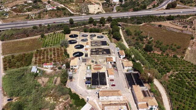 Urban wastewater treatment plant seen from above, featuring circular clarifier tanks, sedimentation basins, industrial buildings, surrounding farmland, and a highway nearby