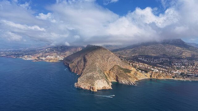 Morro de Toix rocky cape rises above turquoise Mediterranean waters near Calpe, Spain, with coastal town, cliffs and dramatic clouds over mountain ridgeline