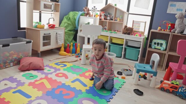 Boy kneeling on colorful foam number mat holding a toy car with outstretched hand amid scattered toys in a playroom; joyful play.