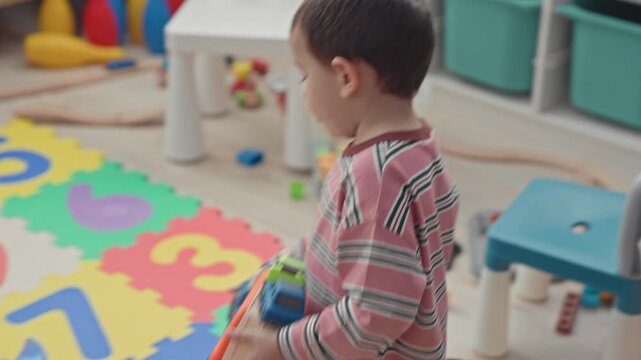 Boy playing with a toy truck on a colorful foam playmat inside a building playroom with storage bins and a white chair; quiet curiosity exploration.