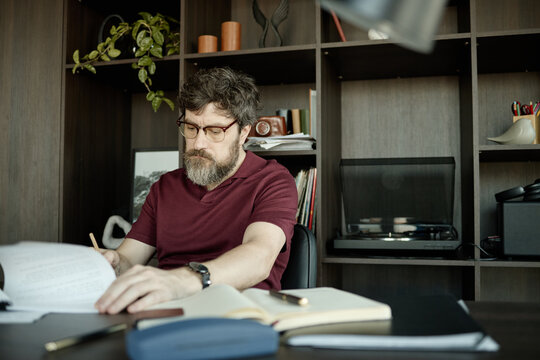 Middle aged Caucasian man reviewing documents and writing notes at desk in home office, managing paperwork and planning tasks

