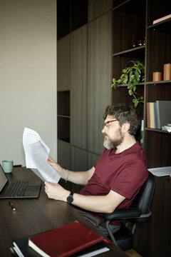 Vertical shot of middle aged Caucasian man reviewing printed chapter of book at desk beside laptop