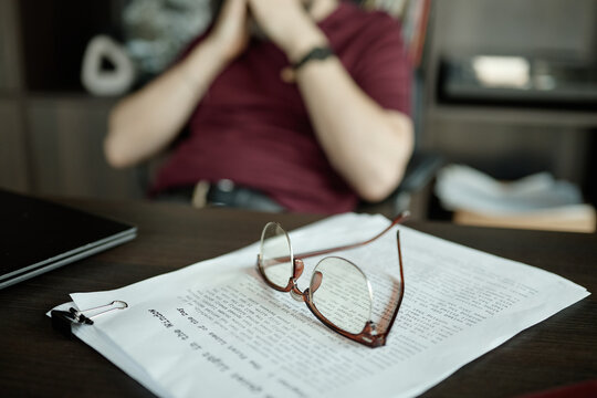 Close up of eyeglasses resting on script pages while middle aged man sitting blurred in background, reviewing document
