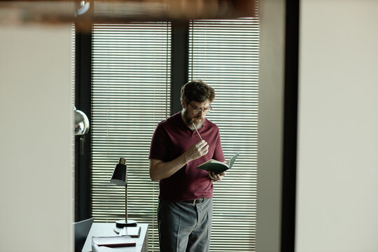 Middle aged Caucasian man reading notebook and thinking in office, planning plot twists and reviewing notes
