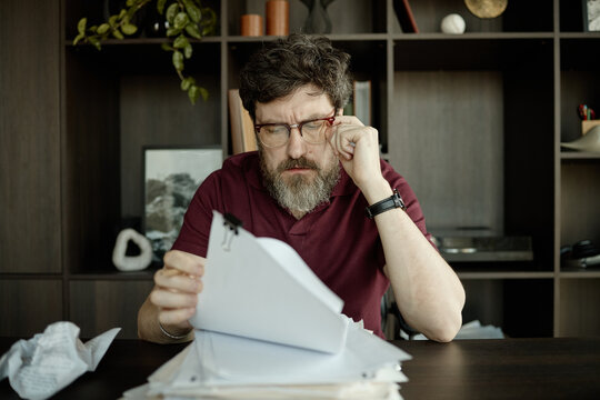 Middle aged Caucasian man reviewing script and touching eyeglasses at desk, showing concentration and stress
