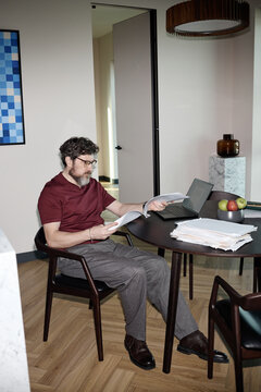 Vertical shot of middle aged Caucasian man reviewing paperwork beside laptop at home office table, managing documents
