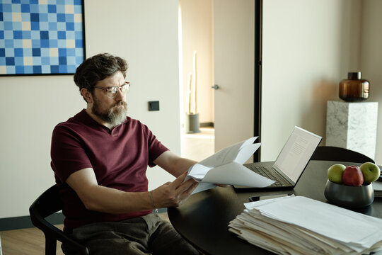 Middle aged Caucasian man reviewing printed script while sitting at dining table in kitchen 
