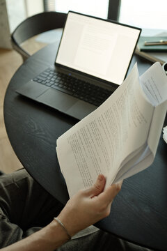Vertical shot of adult man reviewing printed manuscript beside laptop at desk, comparing edits and drafting content
