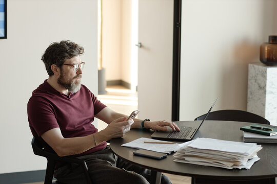 Middle aged Caucasian man working on laptop while checking smartphone at desk, managing multitasking workflow and communication
