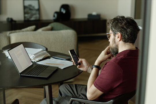Caucasian middle aged man checking smartphone while reviewing laptop chapter of book at home office,
