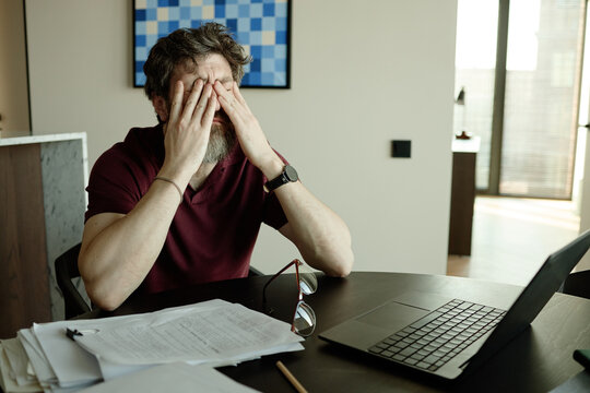 Middle aged Caucasian man covering face while sitting at desk with laptop and paperwork, showing work stress and burnout
