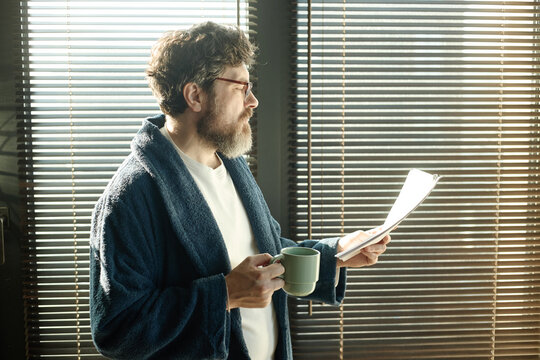 Middle aged Caucasian man reading novel and holding coffee mug by window, reviewing paperwork at home
