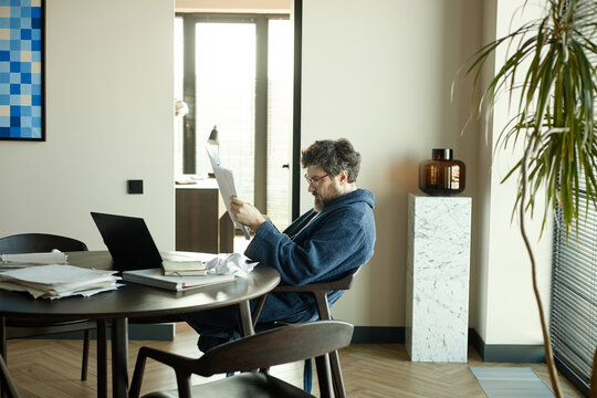 Author dressed in bathrobe sitting at dining table and reading printed chapter of novel 