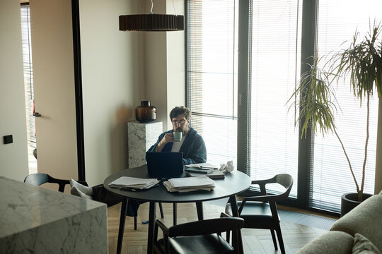 Wide shot of middle aged Caucasian man working on laptop while drinking coffee at home table, managing paperwork
