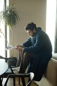 Vertical shot of middle aged Caucasian man reviewing chapter and writing notes at home while working on brand new novel
