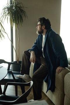 Vertical shot of middle aged Caucasian man sitting on sofa and looking through window at home, reflecting on daily life and solitude
