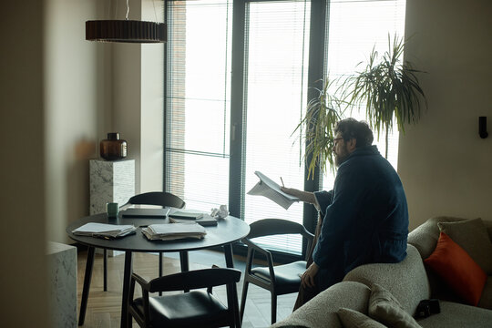 Middle aged Caucasian writer reviewing paperwork at home, checking spelling near dining table
