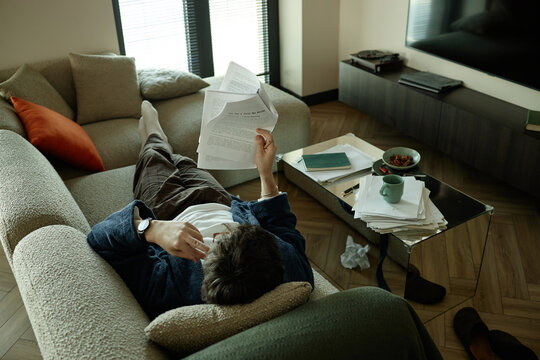 High angle view of young adult man reviewing script pages while reclining on sofa beside scattered drafts and notes, suggesting creative writing workflow
