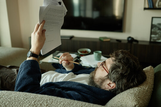 Caucasian young adult man reviewing printed pages of novel while resting on sofa and holding pencil
