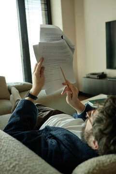 Vertical shot of middle aged Caucasian man reviewing printed script and marking notes with pencil while reclining at home. Useful for remote work, editing, writing, productivity
