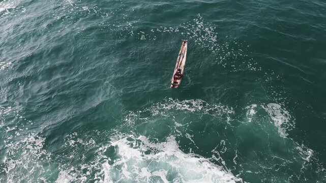 Aerial view of fishermen in a traditional wooden boat navigating the turquoise sea with white foam and gentle waves in Kovalam, Kerala, India.