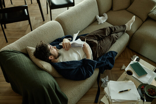 Young adult Caucasian man lying on sofa, reviewing documents and holding pen, showing burnout and work overload at home
