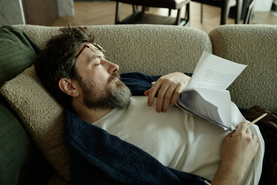 Middle aged Caucasian man resting on sofa while holding paperwork and pencil, suggesting burnout or fatigue
