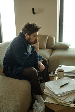Vertical shot of middle aged Caucasian man sitting on sofa, holding glasses and thinking over pages of novel, showing stress and burnout
