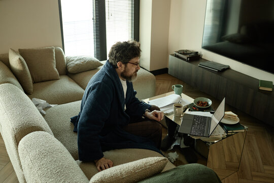 High angle view of middle aged Caucasian man working on laptop from sofa, managing documents and breakfast at home

