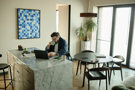 Wide shot of middle aged Caucasian man working on laptop while drinking coffee at kitchen counter in apartment, representing remote work routine
