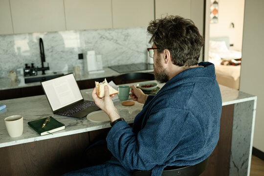 Caucasian adult man eating breakfast and drinking coffee while reading on laptop at kitchen counter, multitasking at home. Ideal for remote work, freelance lifestyle, morning routine
