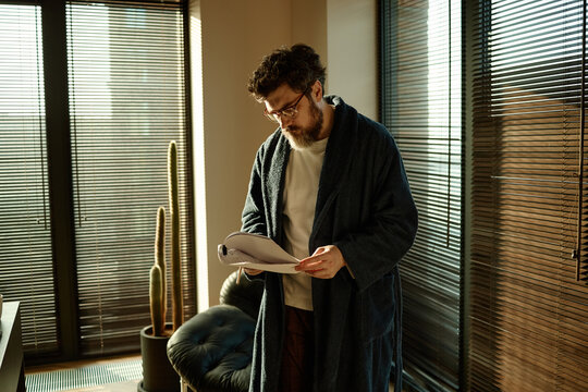 Caucasian author dressed in bathrobe reading chapters of printed novel while working at home office
