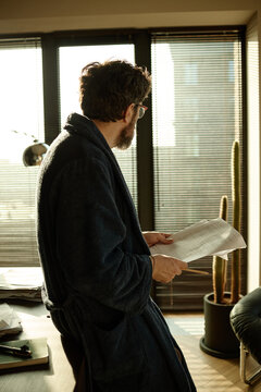 Vertical shot of middle aged Caucasian man reviewing paperwork at home, standing by window and checking documents

