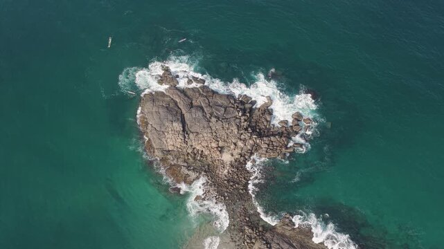 Aerial view of waves crashing against a rocky outcrop in the turquoise ocean with a small boat nearby in Kovalam, Kerala, India.