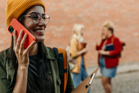 Young woman having phone conversation outdoors near campus wall