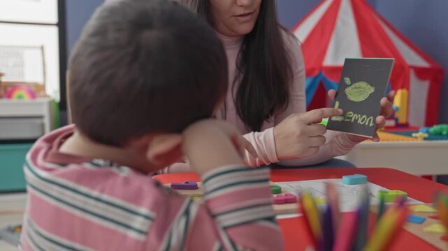Woman teacher holds a lemon flashcard, points finger to the card while guiding a little boy's outstretched hand at a playroom table inside a building; focused learning.