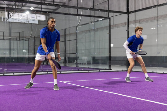 Male teammates standing on purple padel court wearing blue shirts and white shorts holding rackets