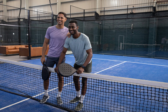 Diverse male friends standing at black mesh net on blue padel court holding rackets near sofas