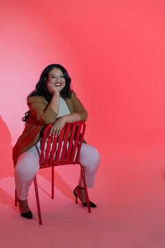 Mid adult female sitting on backward red chair in studio, wearing tan blazer, smiling