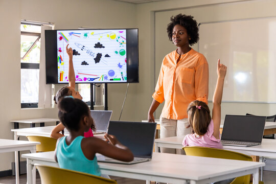 Diverse class teacher wearing orange shirt teaching three kids using laptops in school, copy space