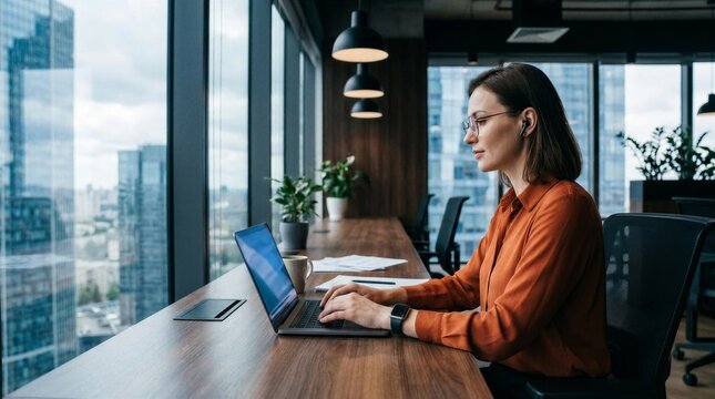 Busy latin hispanic young business woman working on laptop computer sitting at office desk with city view. Indian entrepreneur manager businesswoman using pc for communication, learning at workplace